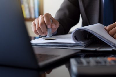 Businessman in suit hand stamping rubber stamp on document in file folder with laptop computer on the desk at office. Authorized allowance permission approval concept.