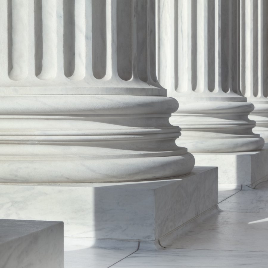 An image of architectural detail from the United States Supreme Court building.  There are circular pillars visible in a row.  The pillars are white.  Each pillar has a block at the bottom, followed by three circular lines going all around them.  Vertical lines come next in an alternating pattern of sunken lines and protruding ones.  There are six pillars visible in the image, but only their lower parts can be seen.  The floor has white tiles.  There is sunlight coming from the front of the building, creating shadows behind the pillars.